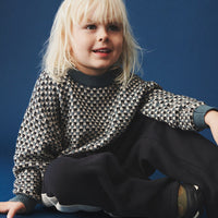 Child wearing a patterned sweater and dark pants sitting on a blue background