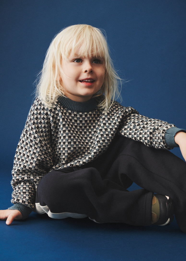 Child wearing a patterned sweater and dark pants sitting on a blue background