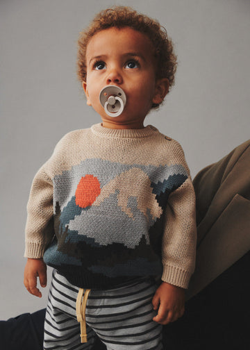 Child wearing a sweater with a nature design and striped pants, holding a pacifier.