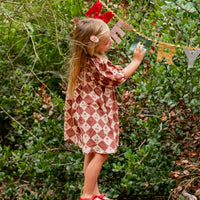 Young girl in a patterned dress standing on a log in a forest, holding a 'Merry' banner.