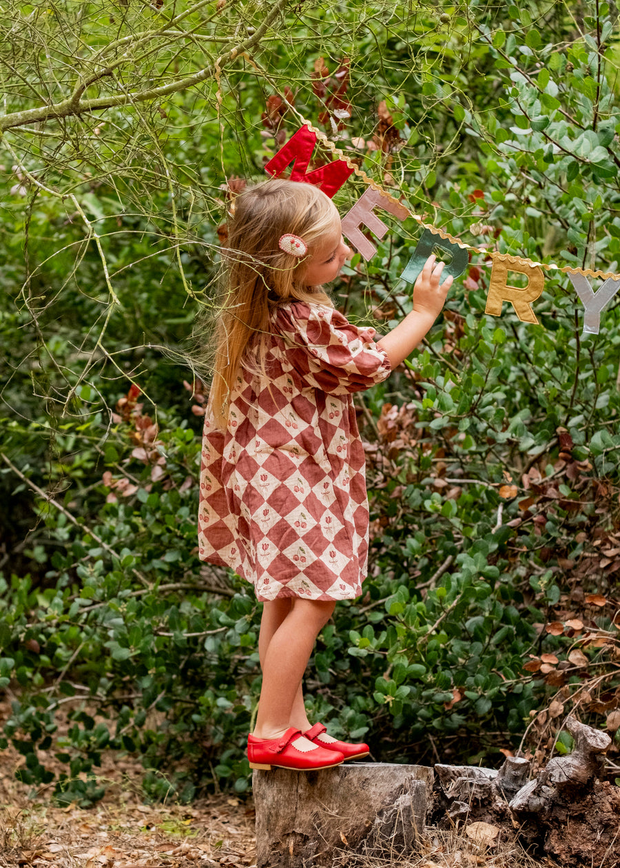 Young girl in a patterned dress standing on a log in a forest, holding a 'Merry' banner.