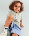 Child wearing a striped shirt and blue shorts sitting on a light gray background