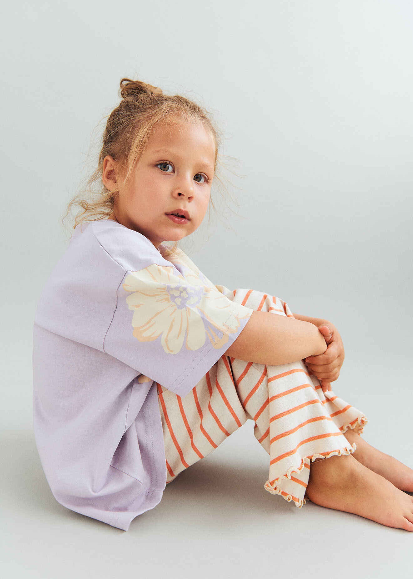 Young girl wearing a light purple shirt with a floral design and striped pants on a plain background