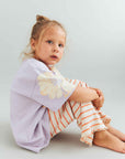 Young girl wearing a light purple shirt with a floral design and striped pants on a plain background
