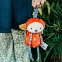 Child holding a small plush toy with an orange outfit and red hat against a natural background.
