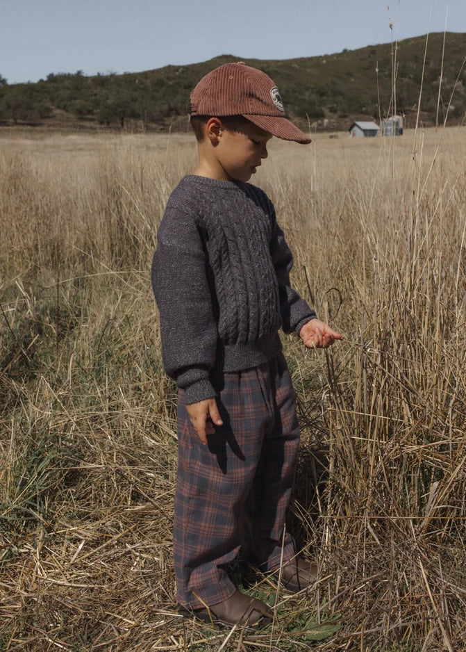 A child standing in a dry grassy field wearing a dark gray cable-knit sweater, plaid pants, and a brown corduroy cap.