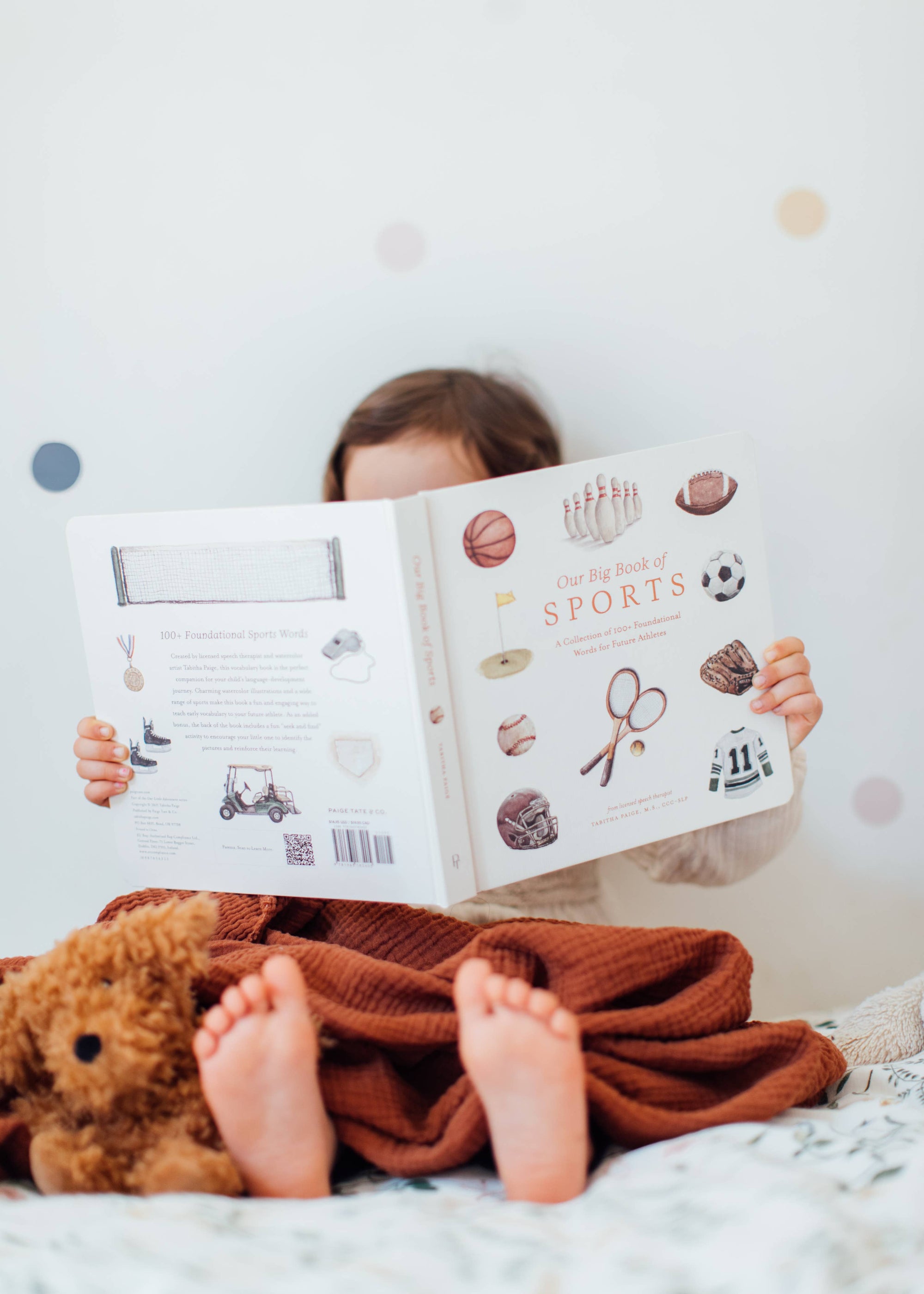 Child reading a book with a teddy bear on a bed