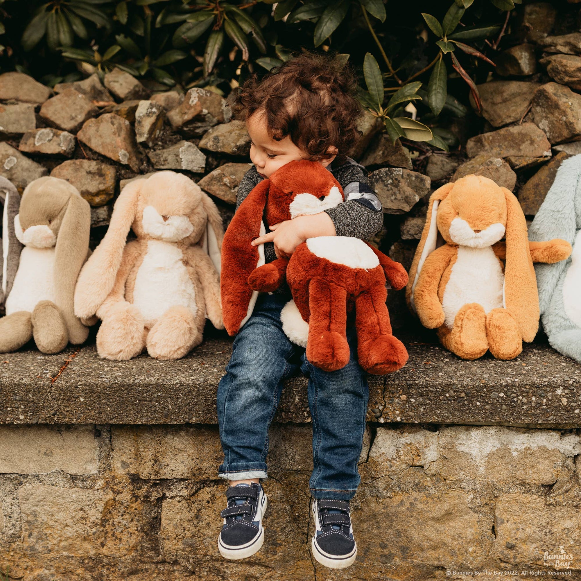 Child holding a plush deer toy with stuffed animals on a stone ledge.