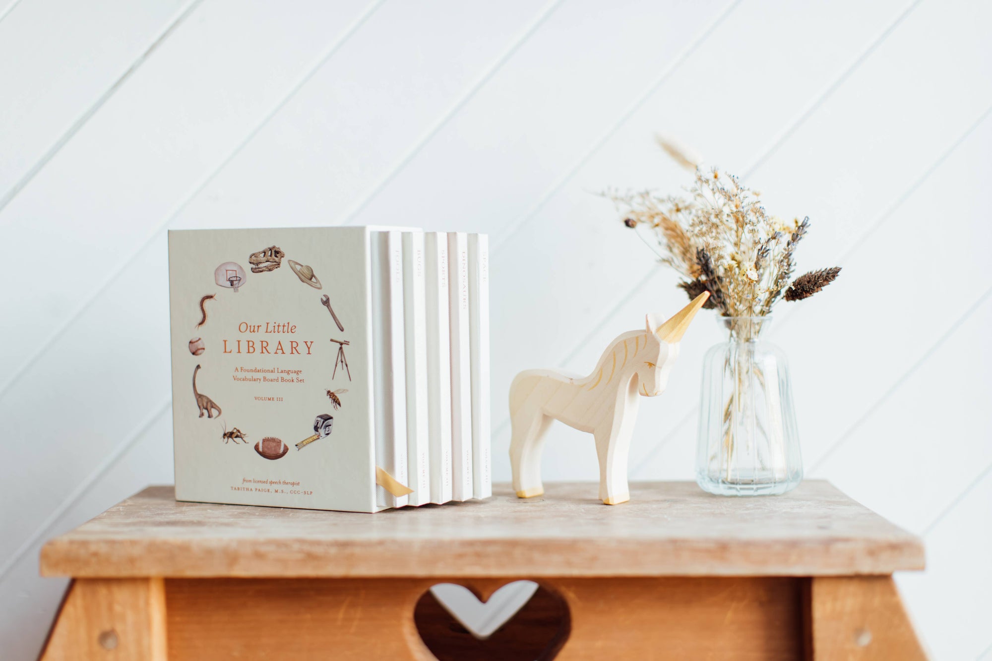 Book titled 'Our Little Library' on a wooden shelf with a unicorn figurine and vase of dried flowers.
