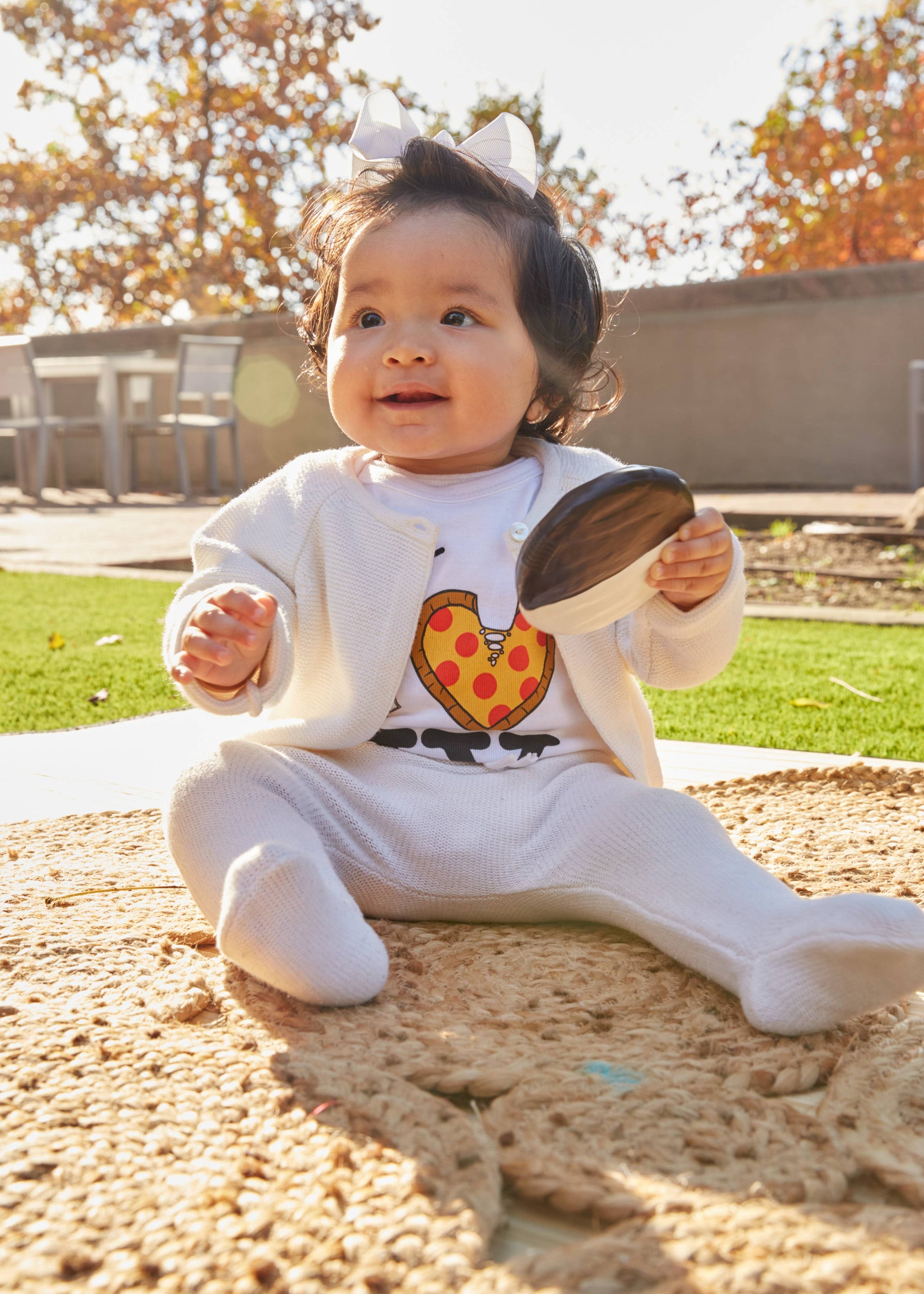 Baby sitting on a playground with a toy, surrounded by trees with autumn leaves.