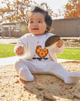 Baby sitting on a playground with a toy, surrounded by trees with autumn leaves.