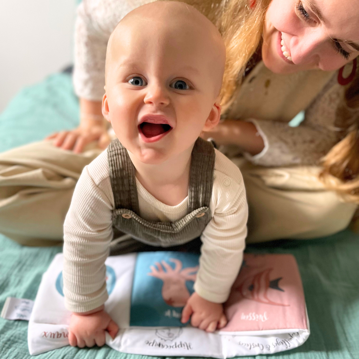 A baby wearing a cream ribbed long-sleeve shirt and brown corduroy overalls crawls on a mint green blanket, holding a baby book. A smiling woman in a cream knit top and beige pants leans over the baby from behind.