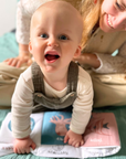 A baby wearing a cream ribbed long-sleeve shirt and brown corduroy overalls crawls on a mint green blanket, holding a baby book. A smiling woman in a cream knit top and beige pants leans over the baby from behind.