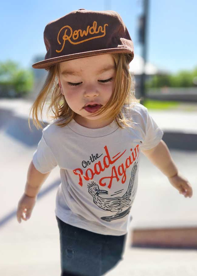 Child wearing a cap and t-shirt with 'On the Road Again' text at a skate park.