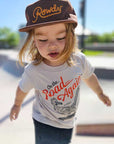 Child wearing a cap and t-shirt with 'On the Road Again' text at a skate park.