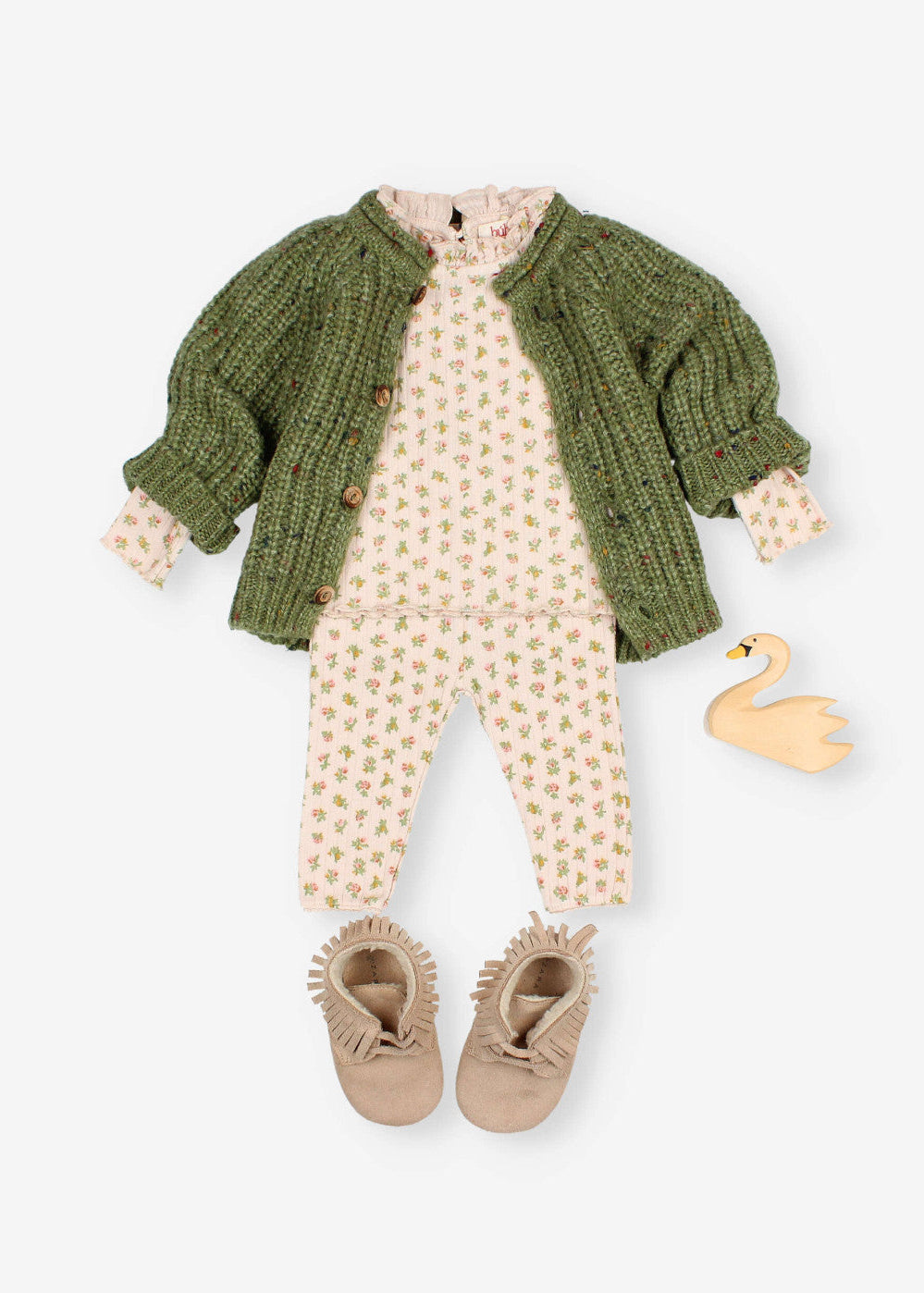 A baby clothing set laid flat on a light gray background, including a green knit cardigan, a cream floral romper, matching pants, tan fringed booties, and a small yellow swan toy.