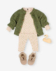 A baby clothing set laid flat on a light gray background, including a green knit cardigan, a cream floral romper, matching pants, tan fringed booties, and a small yellow swan toy.