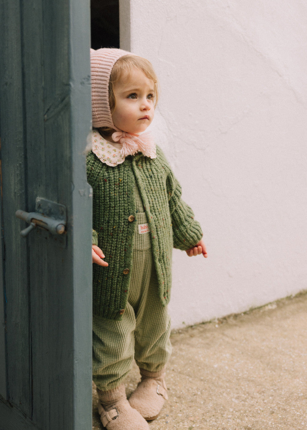 Child wearing a green knitted outfit peeking out from behind a door.