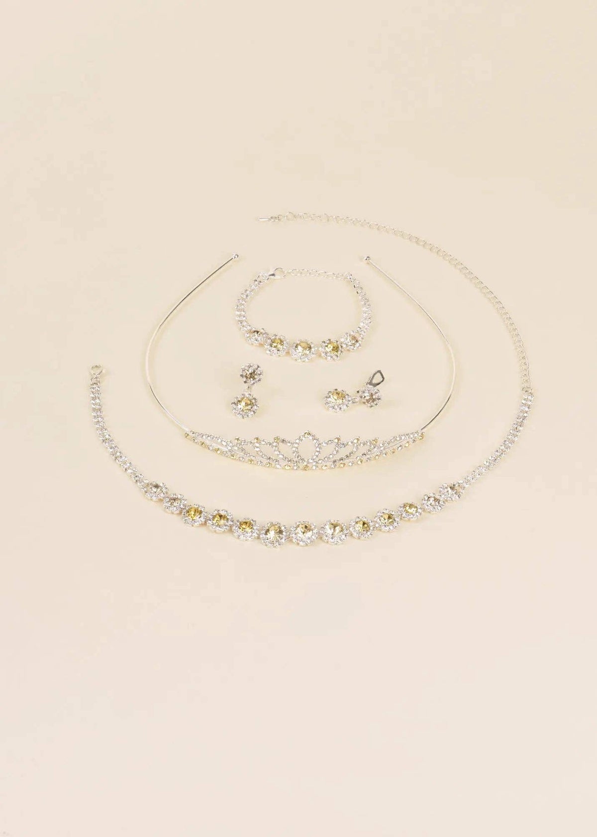 A coordinated jewelry set on a warm cream background, including a silver-tone tiara necklace, matching bracelet, earrings, and a curved collar-style necklace with clear rhinestones.