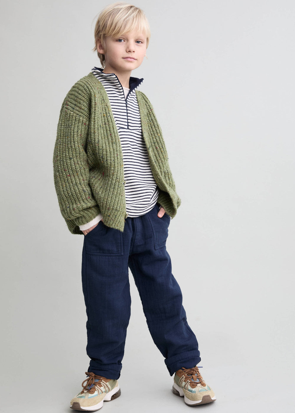 Child wearing a green cardigan, striped shirt, and blue pants on a plain background