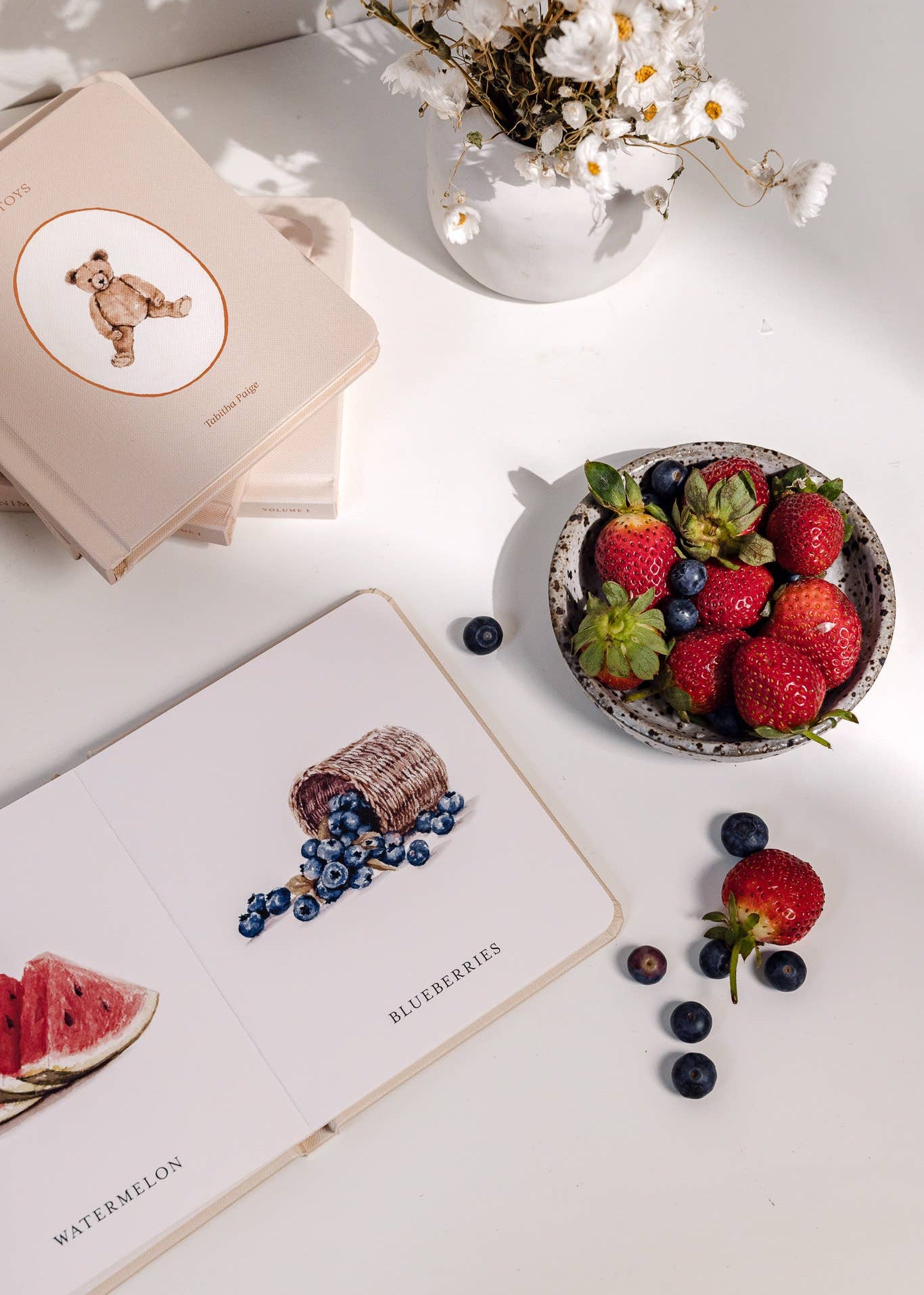 Bowl of strawberries and blueberries on a white surface with books and flowers.