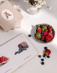 Bowl of strawberries and blueberries on a white surface with books and flowers.