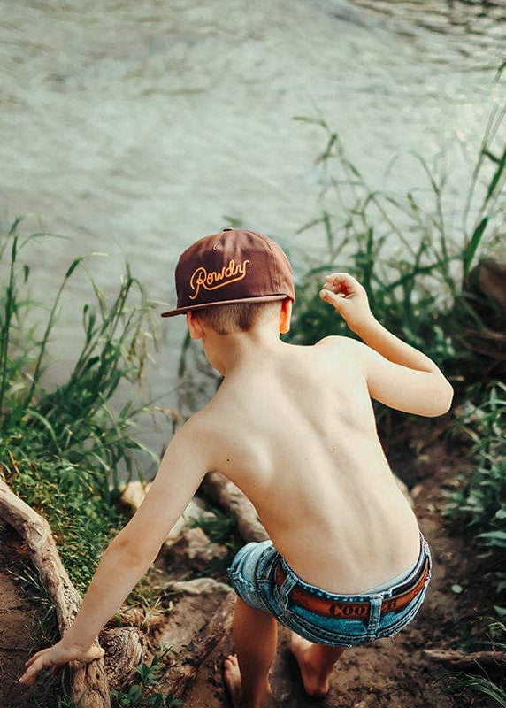 Child wearing a cap and shorts standing near a body of water with greenery.