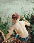 Child wearing a cap and shorts standing near a body of water with greenery.