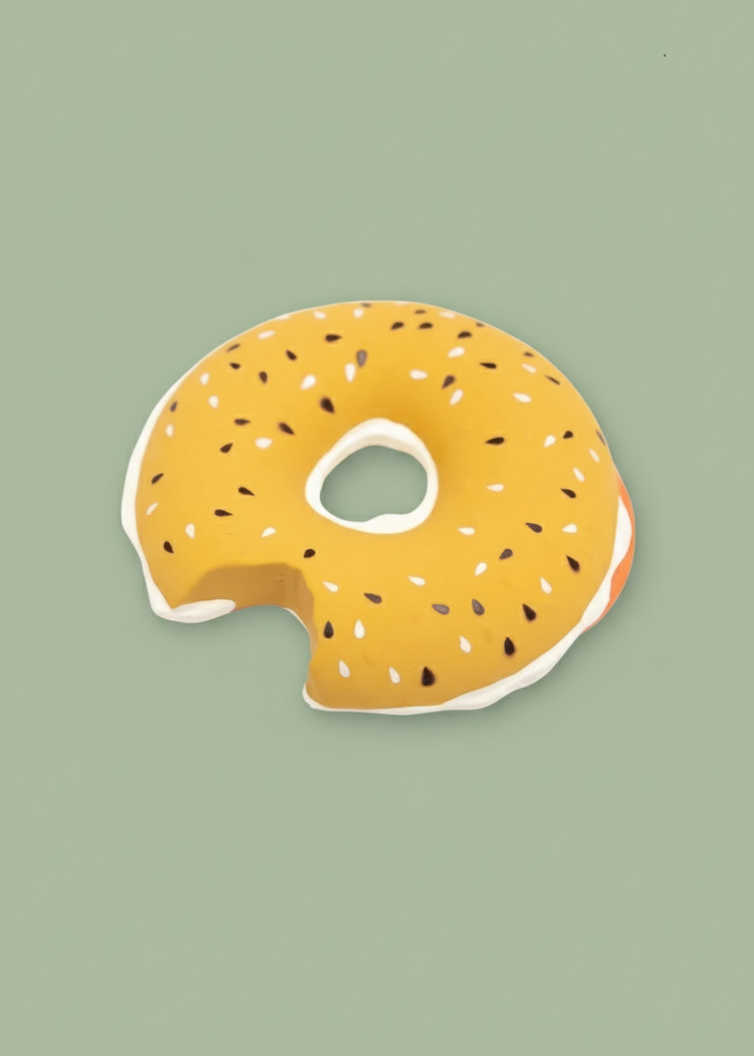 A novelty donut-shaped bath mat or pool float in a mustard yellow glaze with white frosting and black sesame seeds, shown on a solid sage green background.