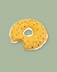 A novelty donut-shaped bath mat or pool float in a mustard yellow glaze with white frosting and black sesame seeds, shown on a solid sage green background.