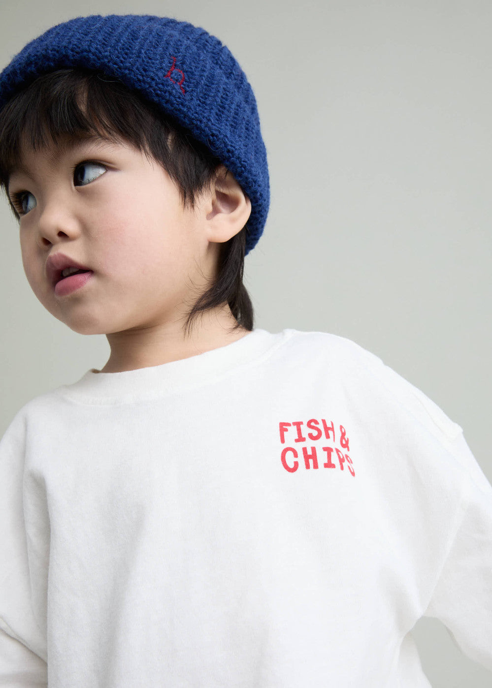 A young child wearing a blue knit beanie and a white sweatshirt with red 'FISH & CHIPS' text on the chest, photographed against a plain light gray background.