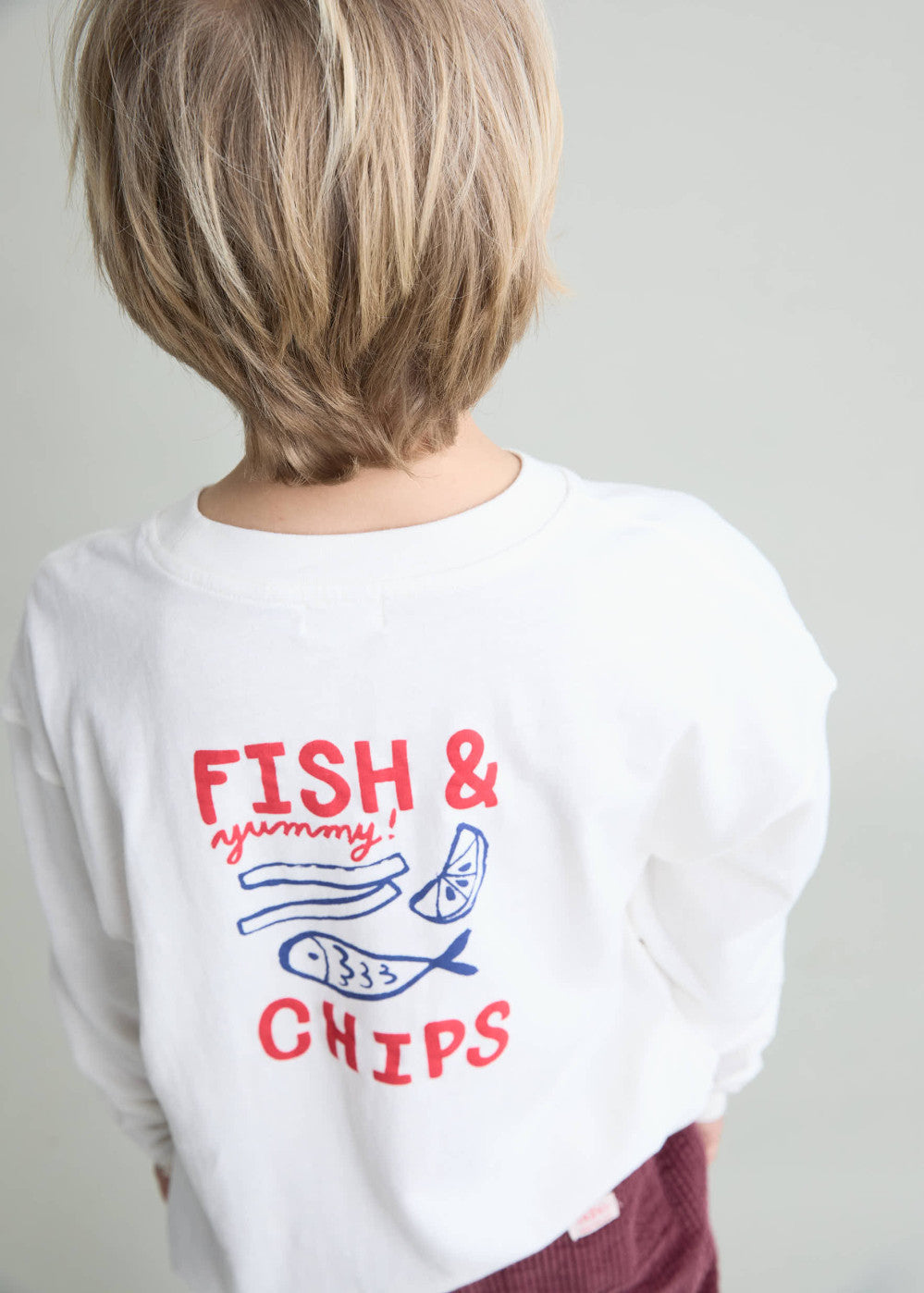 A child shown from behind wearing a white long-sleeve t-shirt with a red and blue graphic print that reads 'FISH & yummy! CHIPS' with illustrations of a fish and chips, set against a plain light gray background.