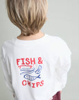 A child shown from behind wearing a white long-sleeve t-shirt with a red and blue graphic print that reads 'FISH & yummy! CHIPS' with illustrations of a fish and chips, set against a plain light gray background.