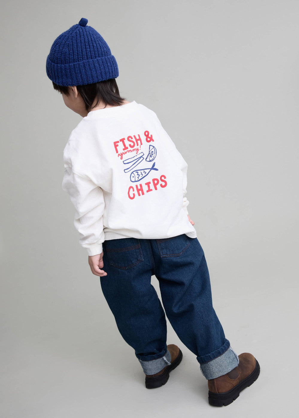 A child shown from the back wearing a white sweatshirt with a red and blue 'FISH & CHIPS' graphic, dark blue jeans, and a navy blue knit beanie, standing against a light gray studio background.