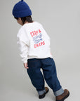 A child shown from the back wearing a white sweatshirt with a red and blue 'FISH & CHIPS' graphic, dark blue jeans, and a navy blue knit beanie, standing against a light gray studio background.
