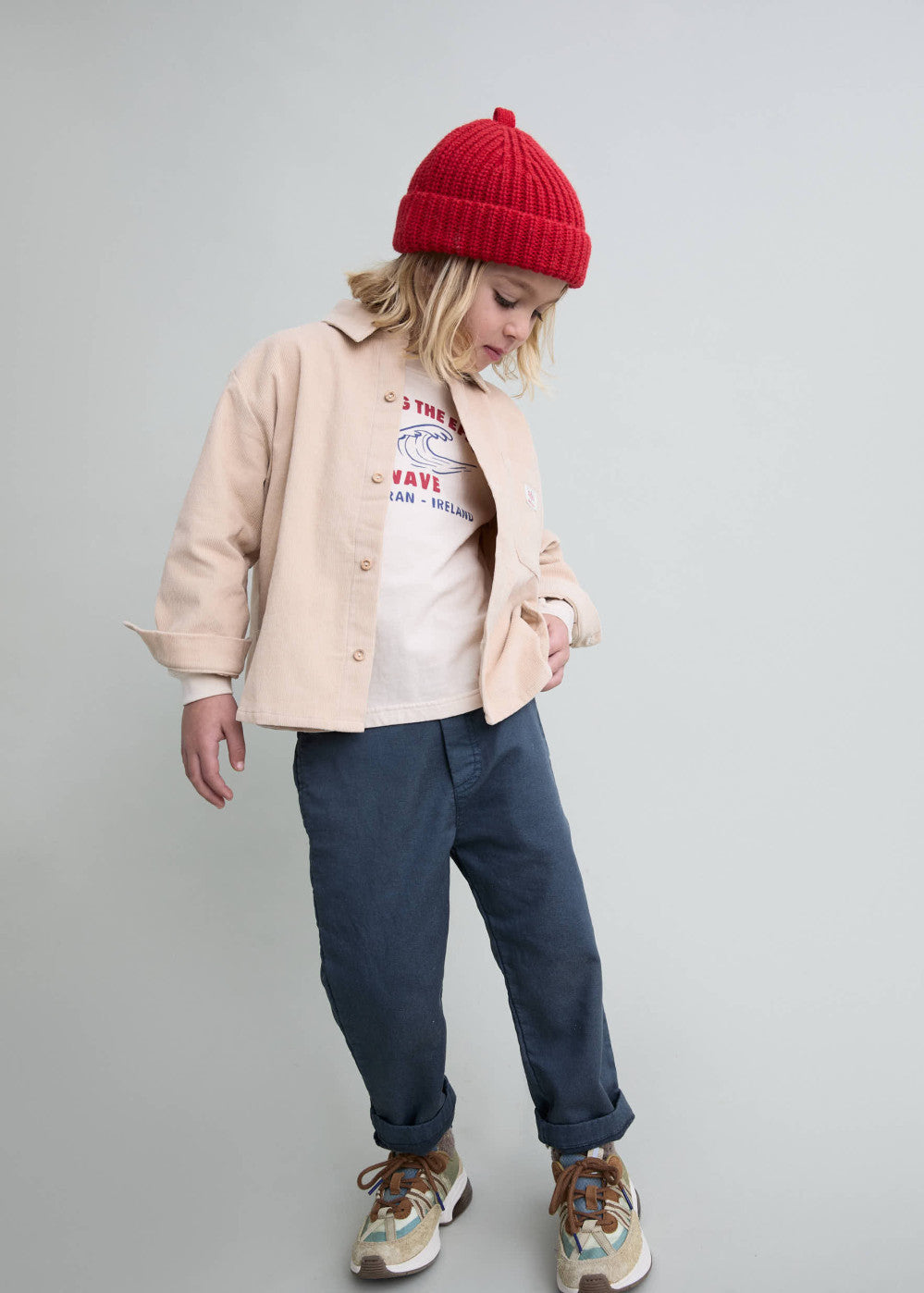 A child model wearing a light beige button-up overshirt, a white graphic t-shirt, navy blue pants, a red knit beanie, and sneakers, standing against a light gray studio background.
