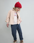 A child model wearing a light beige button-up overshirt, a white graphic t-shirt, navy blue pants, a red knit beanie, and sneakers, standing against a light gray studio background.