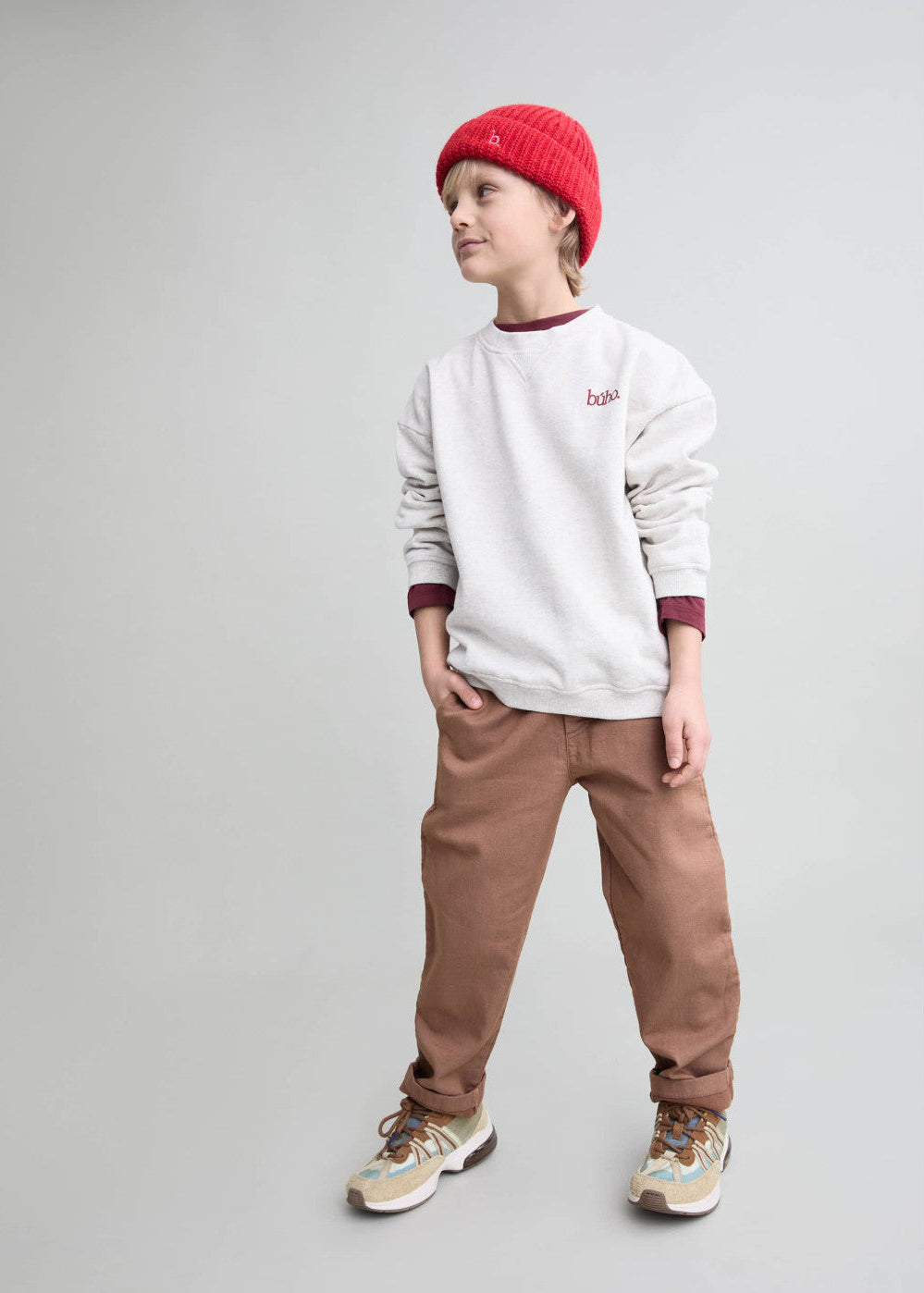 A child model wearing a white crewneck sweatshirt with burgundy trim, brown pants, a red knit beanie, and patterned sneakers, standing against a solid light gray background.