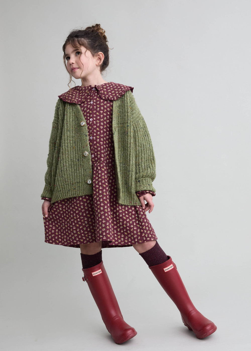 Child wearing a green cardigan, patterned dress, and red boots on a plain background