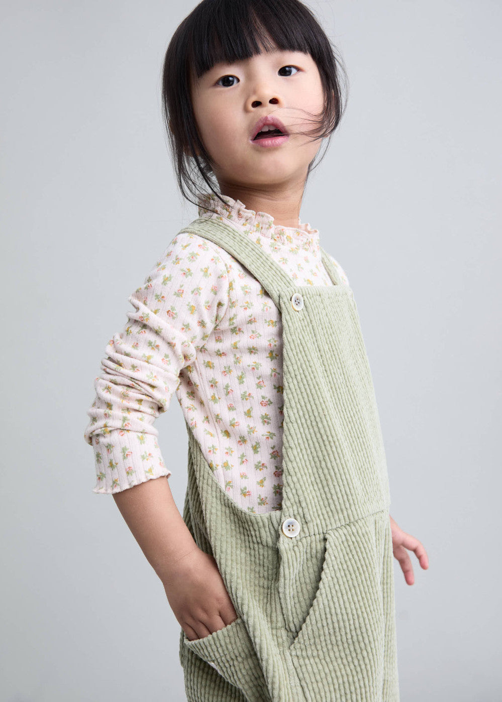 A young child wearing a light sage green corduroy pinafore over a cream floral long-sleeve shirt, photographed against a solid light gray background.
