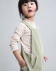 A young child wearing a light sage green corduroy pinafore over a cream floral long-sleeve shirt, photographed against a solid light gray background.