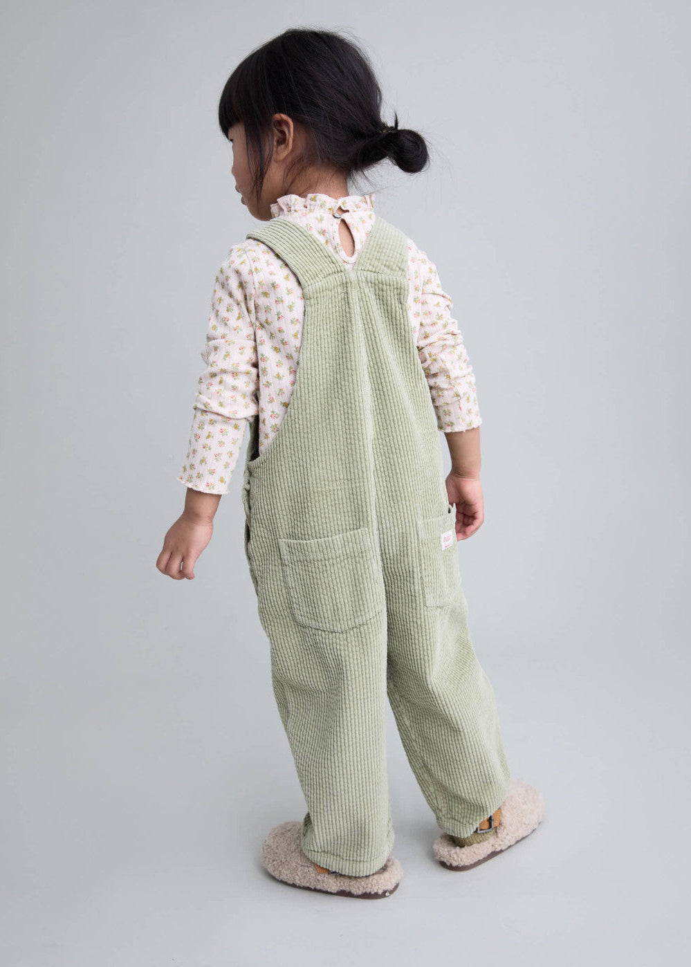 A toddler girl is shown from the back wearing sage green corduroy overall pants over a cream floral long-sleeve top, standing against a solid light gray studio background.