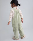 A toddler girl is shown from the back wearing sage green corduroy overall pants over a cream floral long-sleeve top, standing against a solid light gray studio background.
