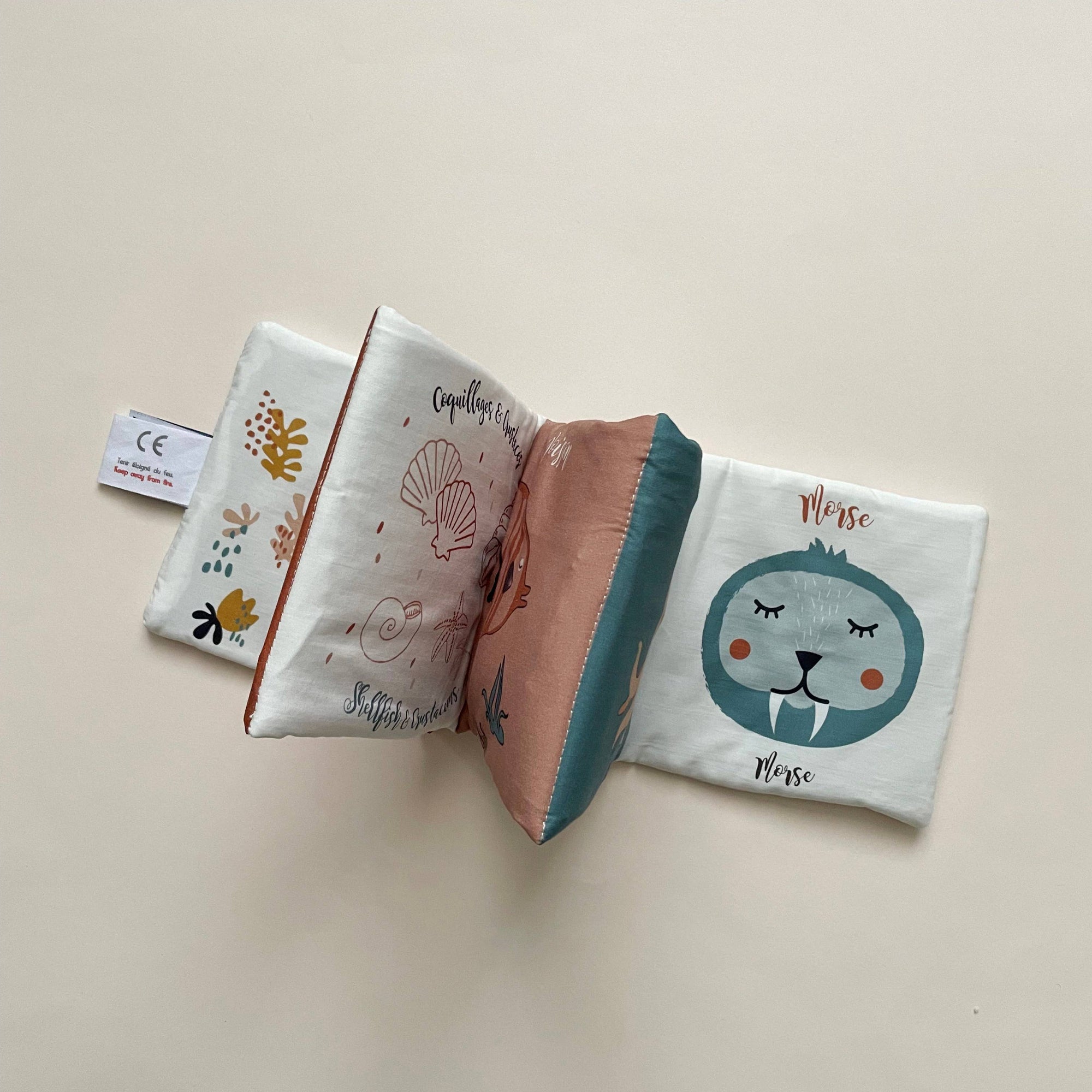 A soft fabric baby book laid flat on a light beige background, showing printed pages with a cartoon animal face, shells, and small illustrations. No person, animal, or real environment is present, so this is Product/Packaging.
