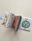 A soft fabric baby book laid flat on a light beige background, showing printed pages with a cartoon animal face, shells, and small illustrations. No person, animal, or real environment is present, so this is Product/Packaging.