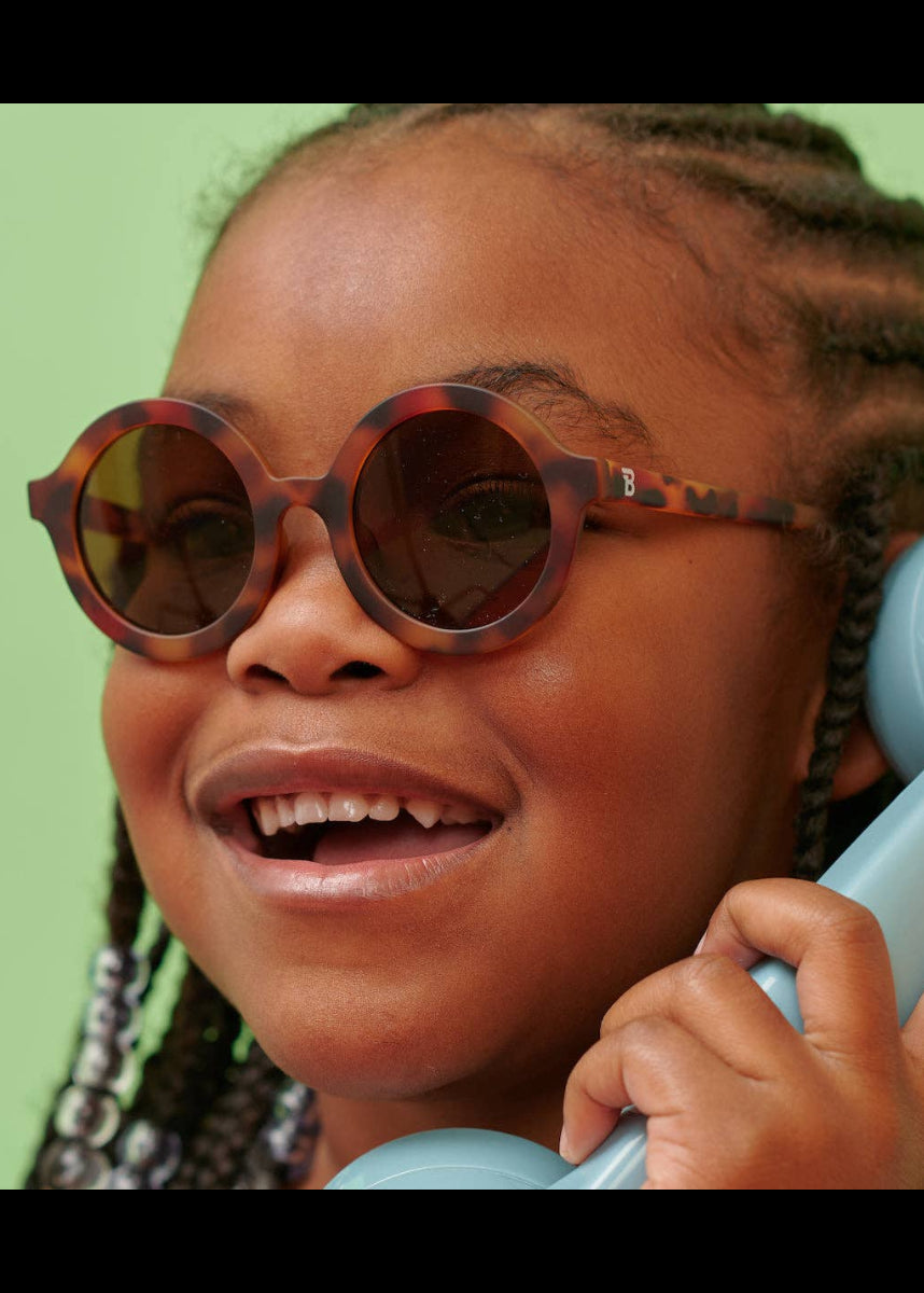 Child wearing tortoiseshell sunglasses and holding a blue phone against a green background