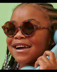 Child wearing tortoiseshell sunglasses and holding a blue phone against a green background