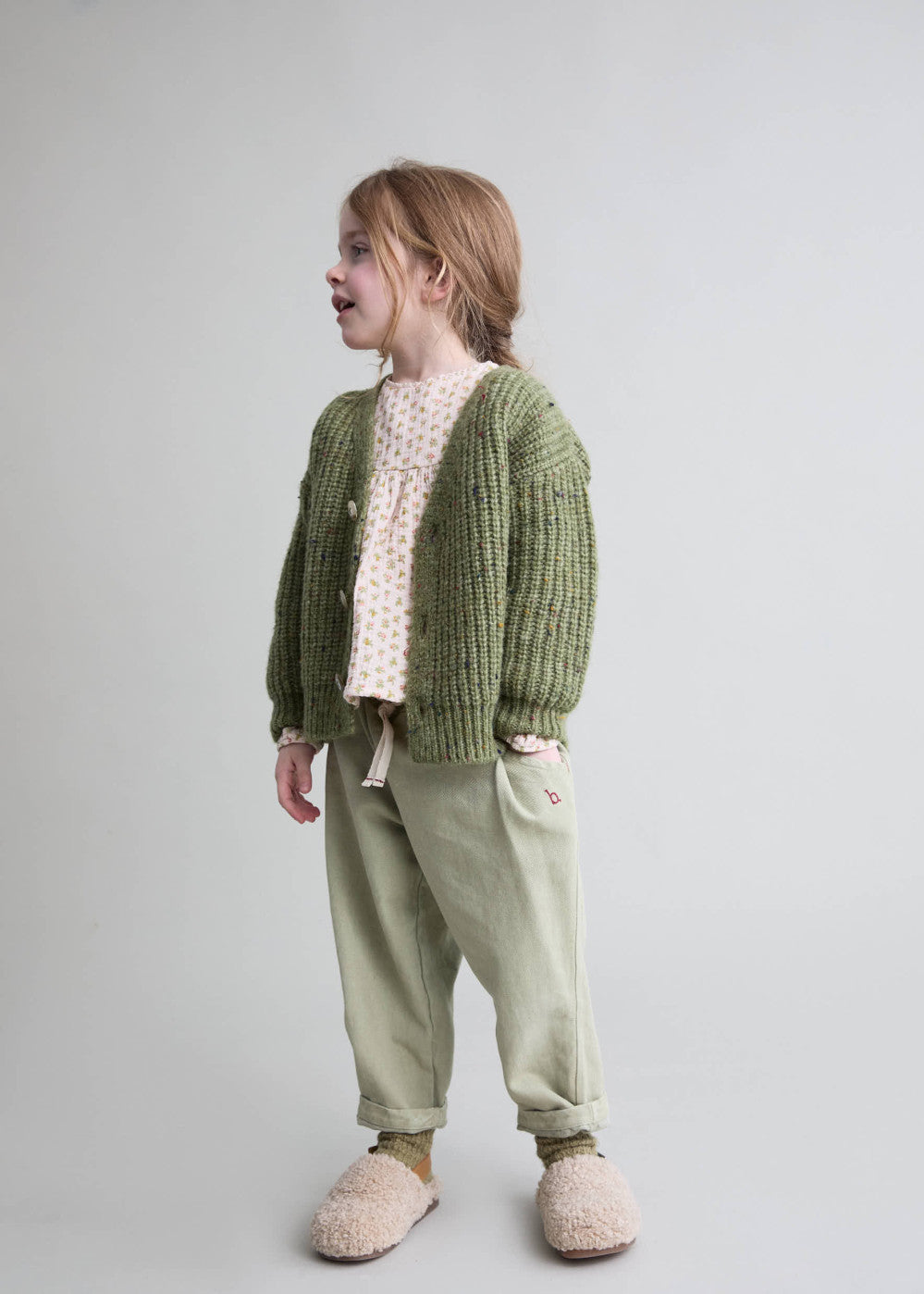 A young girl wearing a green chunky knit cardigan over a cream polka dot top, sage green pants, and fuzzy beige slippers, standing against a light gray studio background.