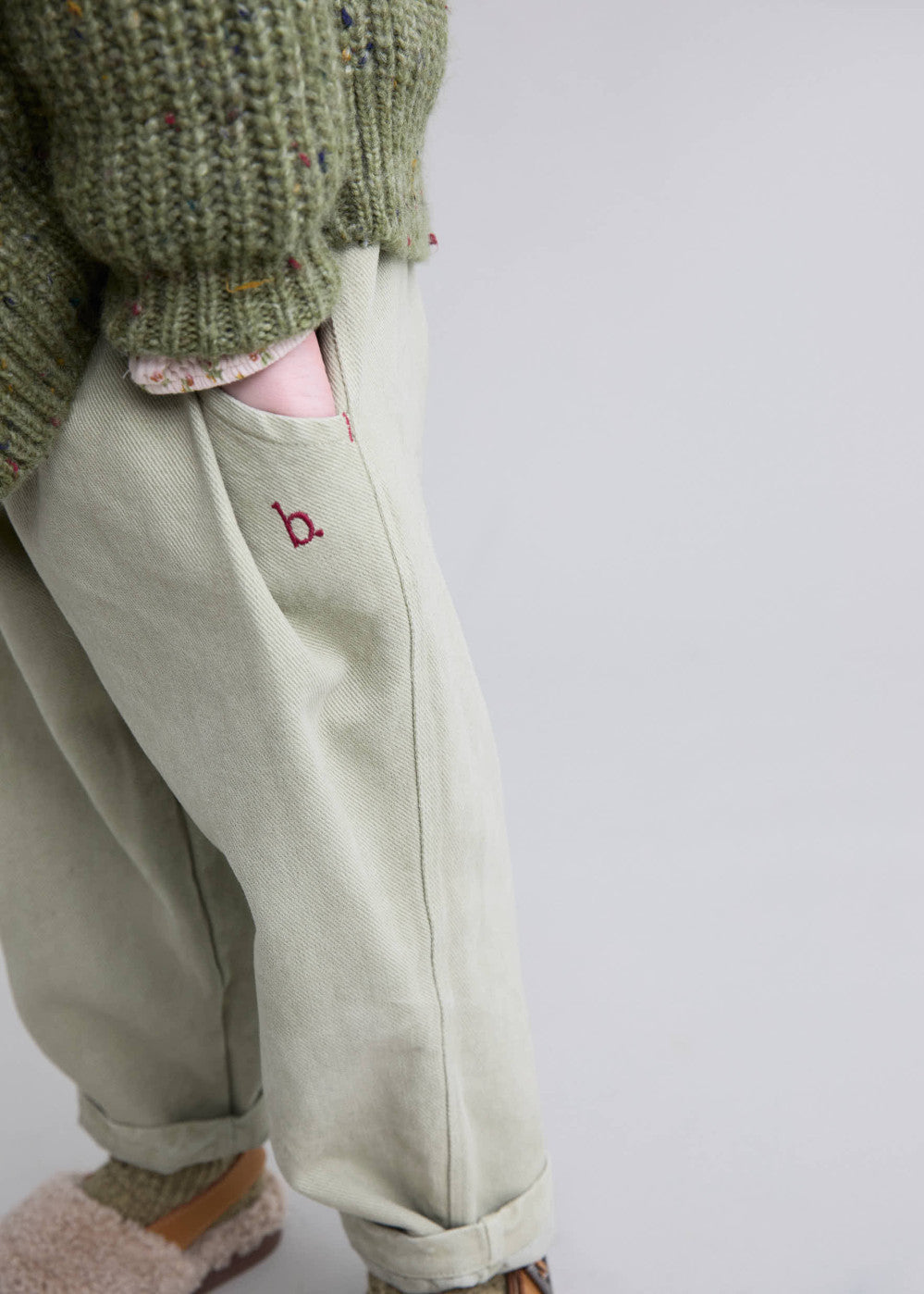 Light-colored pants with a small red logo worn by a person in a green textured sweater on a light gray background
