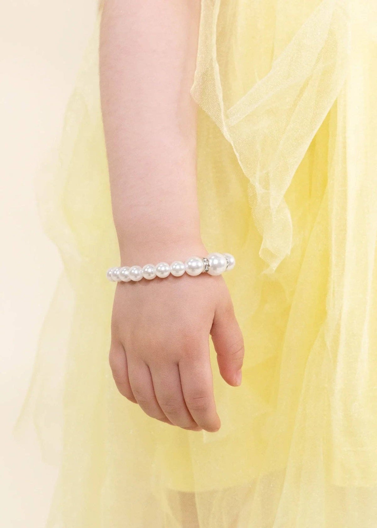 A close-up of a child's hand and wrist wearing a beaded bracelet made of white faux pearls, styled with a soft yellow tulle dress against a light cream background.
